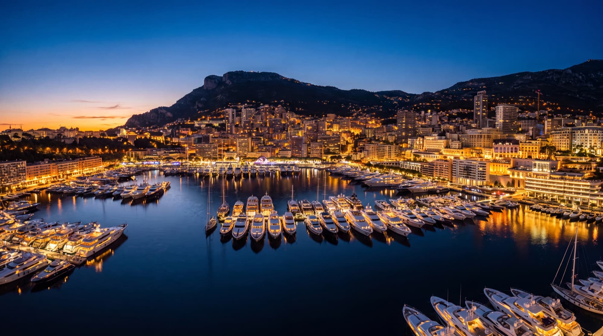 Panorama du Port Hercule à Monaco au crépuscule, superyachts illuminés et Monte-Carlo en arrière-plan